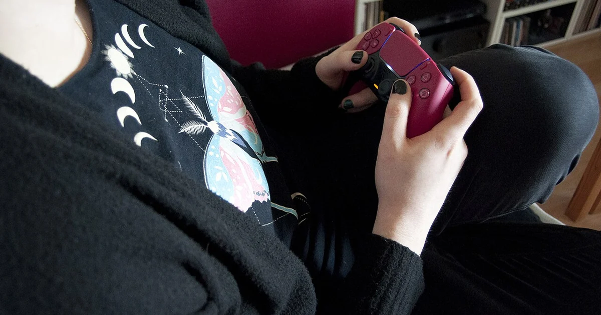 A person sitting is holding a pink PlayStation 5 controller with both hands. Their body is oriented to the right. They are dressed in black, and their T-shirt is printed with a colorful butterfly. Their thumbs are on the analog sticks and t
